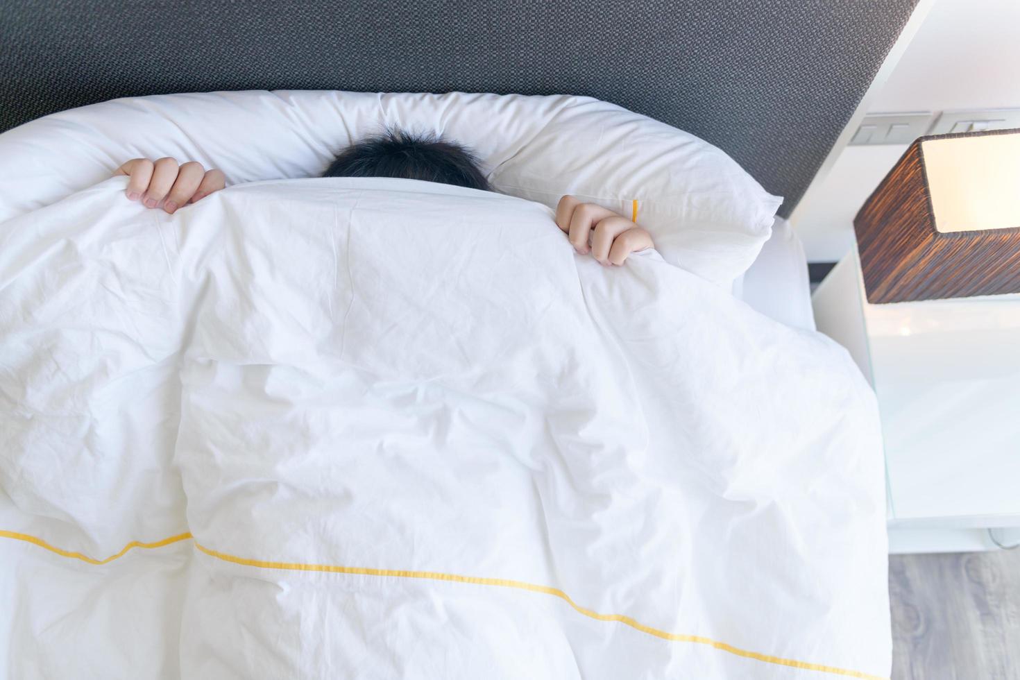 Lazy boy covering his face with blanket while lying in bed at home, photo
