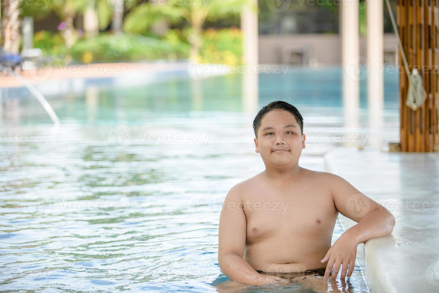 Happy obese boy sit on chair at pool bar in swimming pool, photo