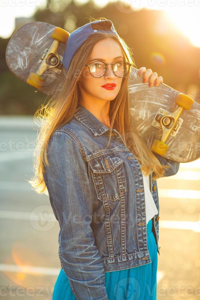Outdoor fashion closeup summer portrait of pretty young woman posing with skateboard in urban youth style in town. Summer evening sunlight photo