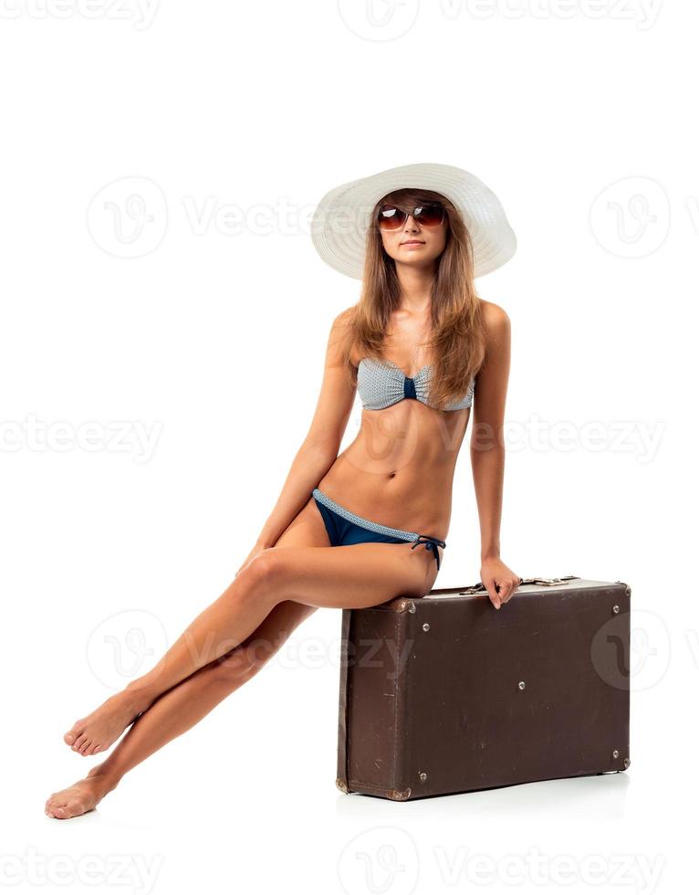 Full length portrait of a beautiful young woman posing in a bikini sitting on a suitcase on a white photo