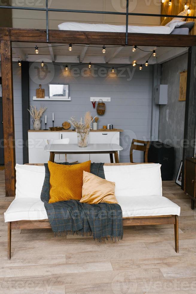 Stylish and Scandinavian interior of the living room of a modern apartment with a white sofa, pillows, a blanket, a kitchen in the background. Modern home decor. photo