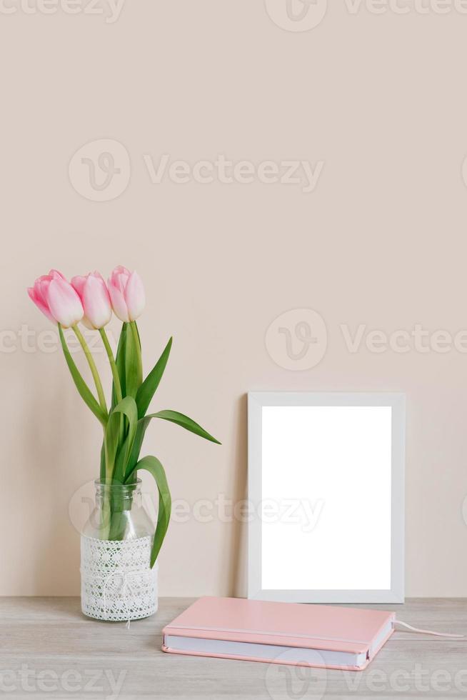 Home interior with decorative elements. Layout with a white frame and pink tulips in a vase and a pink notebook on the table on a light beige background photo