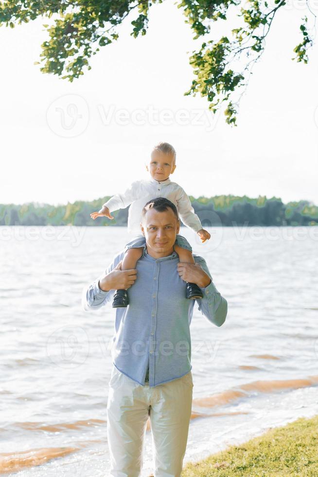 el pequeño hijo es sentado en de papá cuello. el familia camina por el lago en verano foto