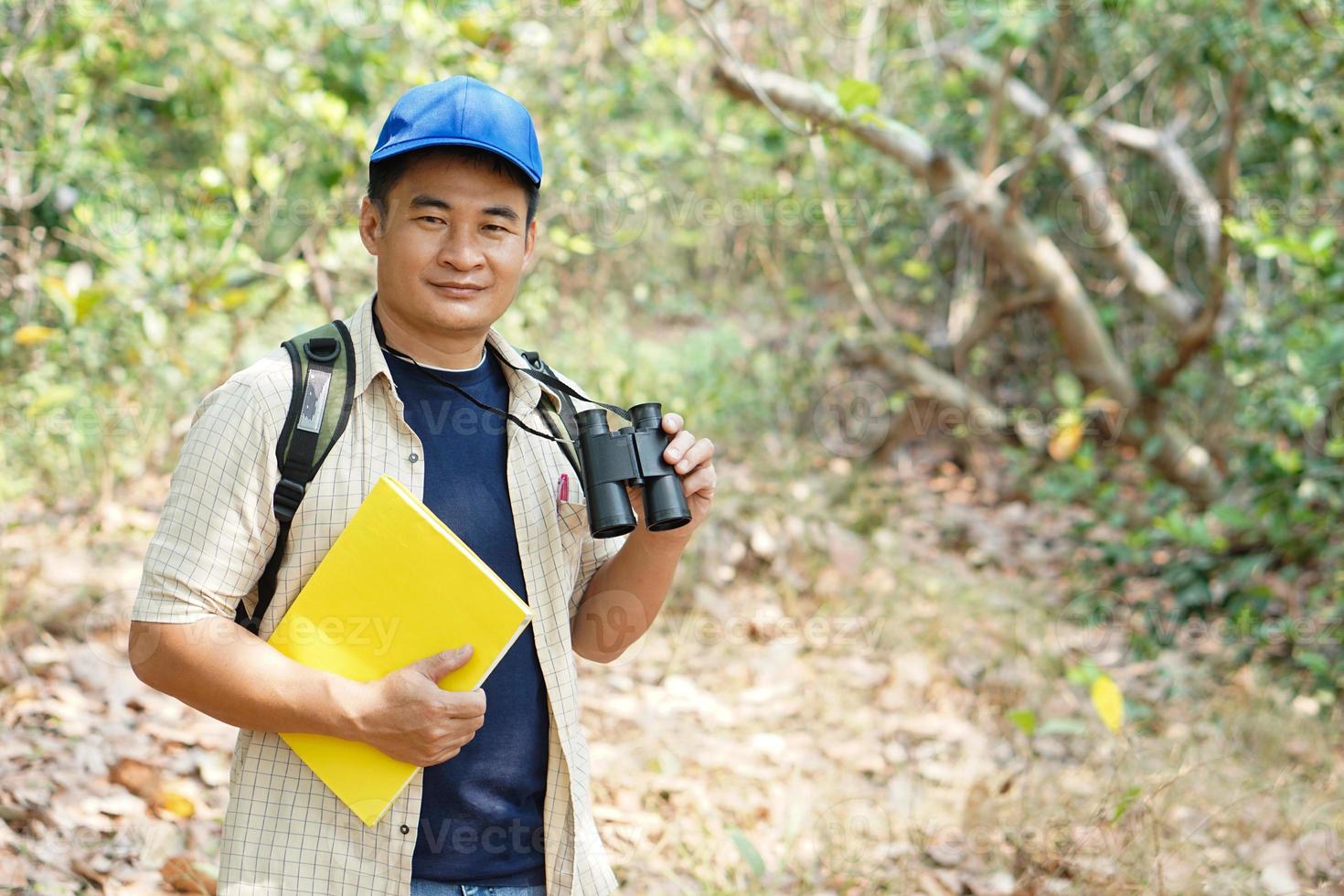 Asian man explorer wears blue cap, holds binocular in forest to survey botanical plants and creatures wildlife. Concept, nature exploration. Ecology and Environment. photo