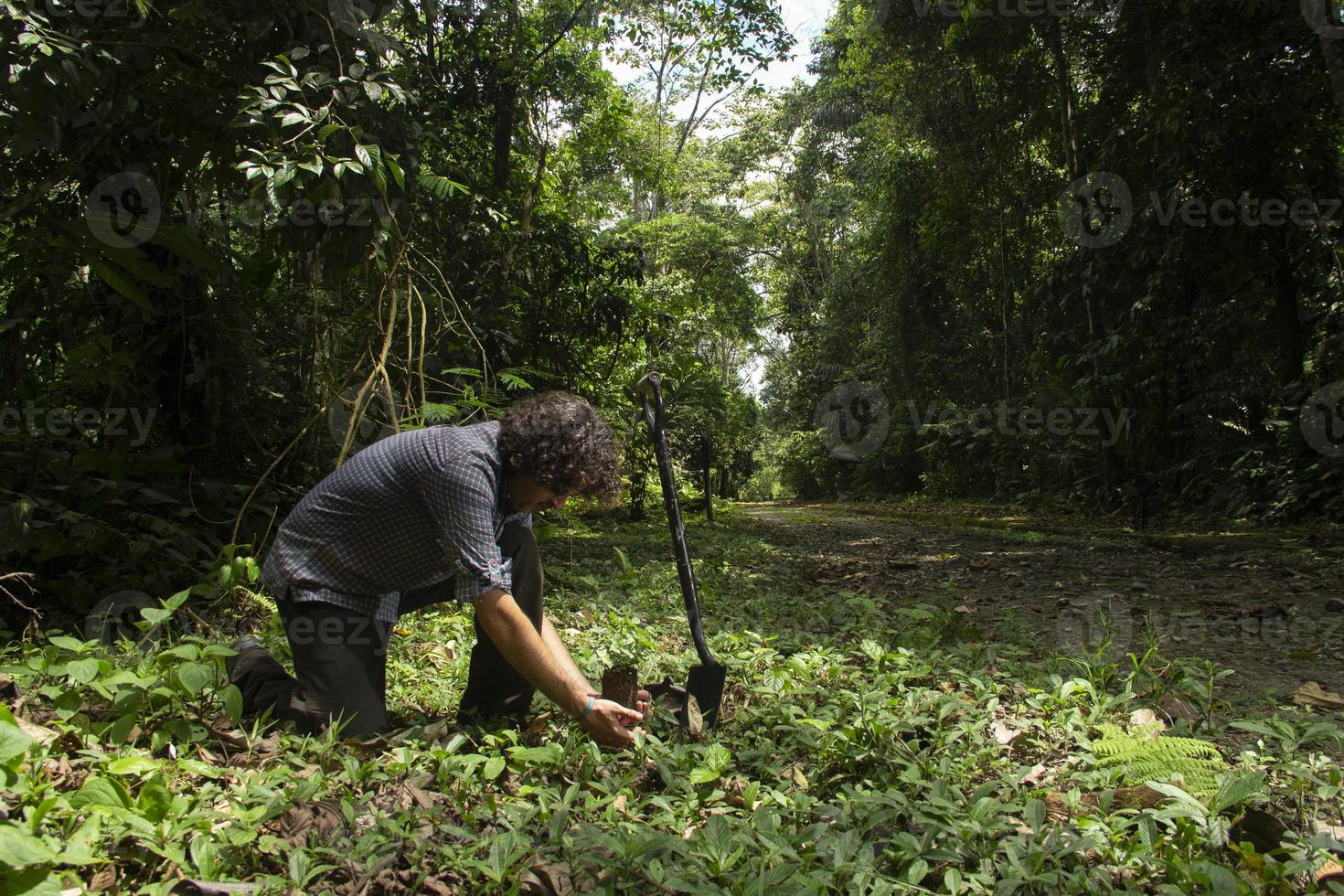 Hispanic man on his knees planting a small plant with a black shovel in a green field surrounded by trees photo