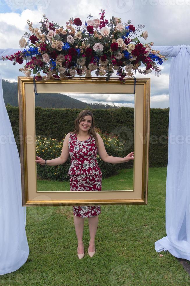 Young latin woman wearing a red dress, standing in a garden posing behind an antique gold frame adorned with a bouquet of roses photo