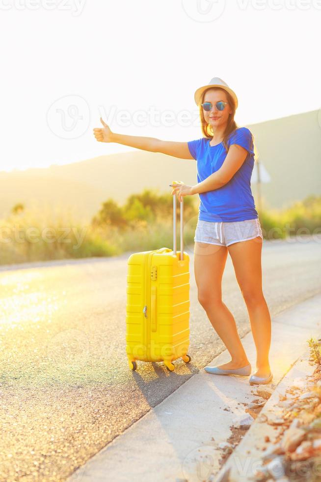 joven mujer con un pulgar arriba y amarillo maleta es de viaje en el la carretera autoestop foto