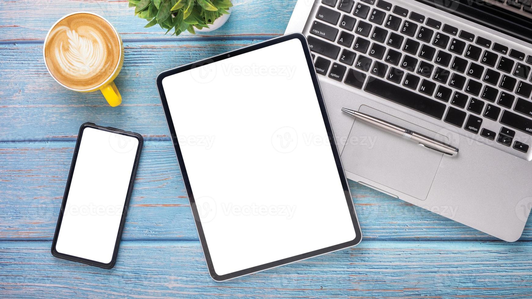 de madera escritorio lugar de trabajo con blanco pantalla tableta y inteligente teléfono, ordenador portátil computadora, bolígrafo y taza de café, parte superior ver con Copiar espacio. foto