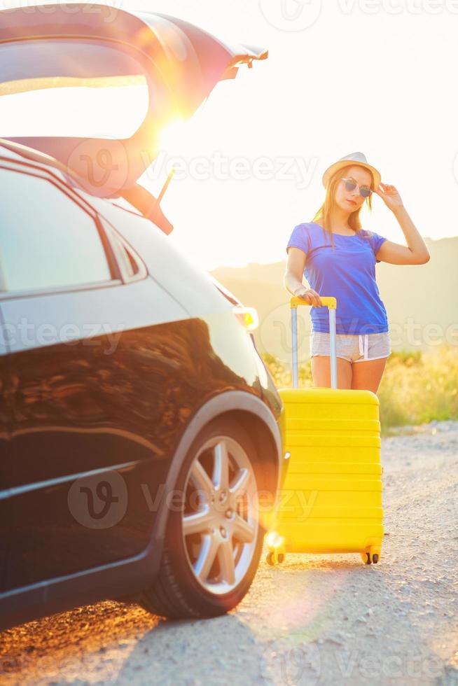 Woman with a yellow suitcase standing near the trunk of a car parked on the roadside photo