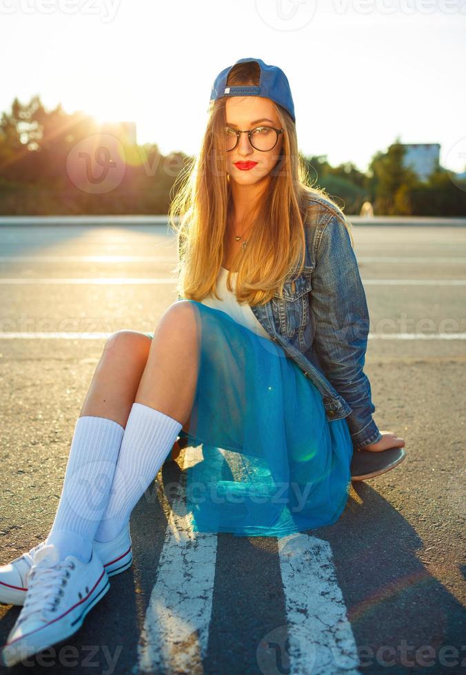 Outdoor fashion closeup summer portrait of pretty young woman posing with skateboard in urban youth style in town. Summer evening sunlight photo