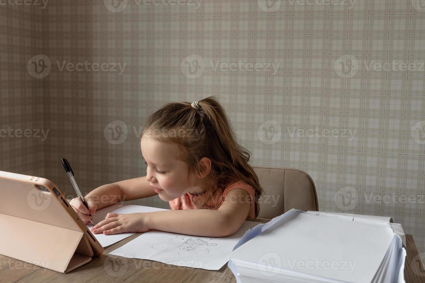 un pequeño niña se sienta a un mesa en frente de su un tableta y un carpeta de blanco papel, el niño sorteos y copias desde el tableta, en línea dibujo lecciones foto