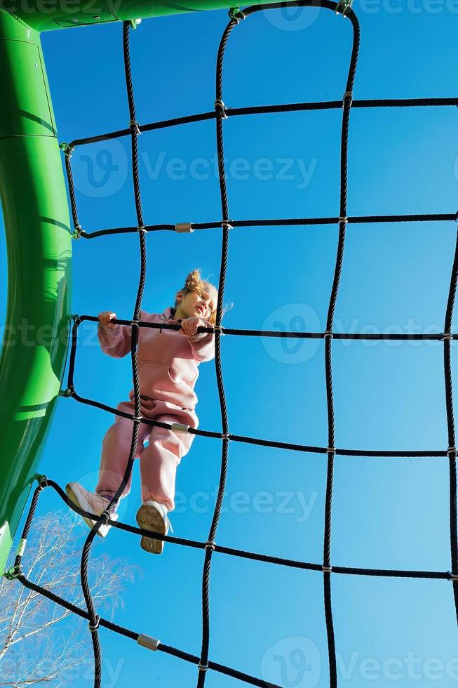 Happy little active girl climbed on a rope web on a playground against a bright blue sky on a sunny day photo