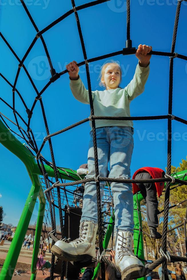 un contento activo diez año antiguo niña subido un cuerda web en un patio de recreo en contra un brillante azul cielo en un soleado día foto