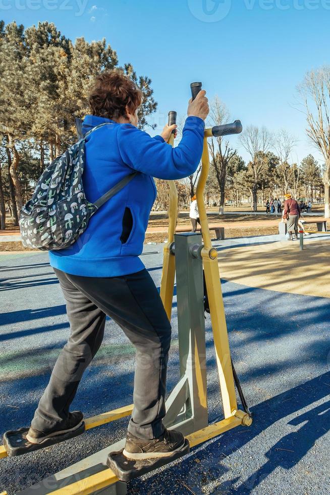 A retired woman with a backpack on her shoulders on an active walk outdoors is engaged in exercise equipment in the park. Senior woman taking care of her health photo