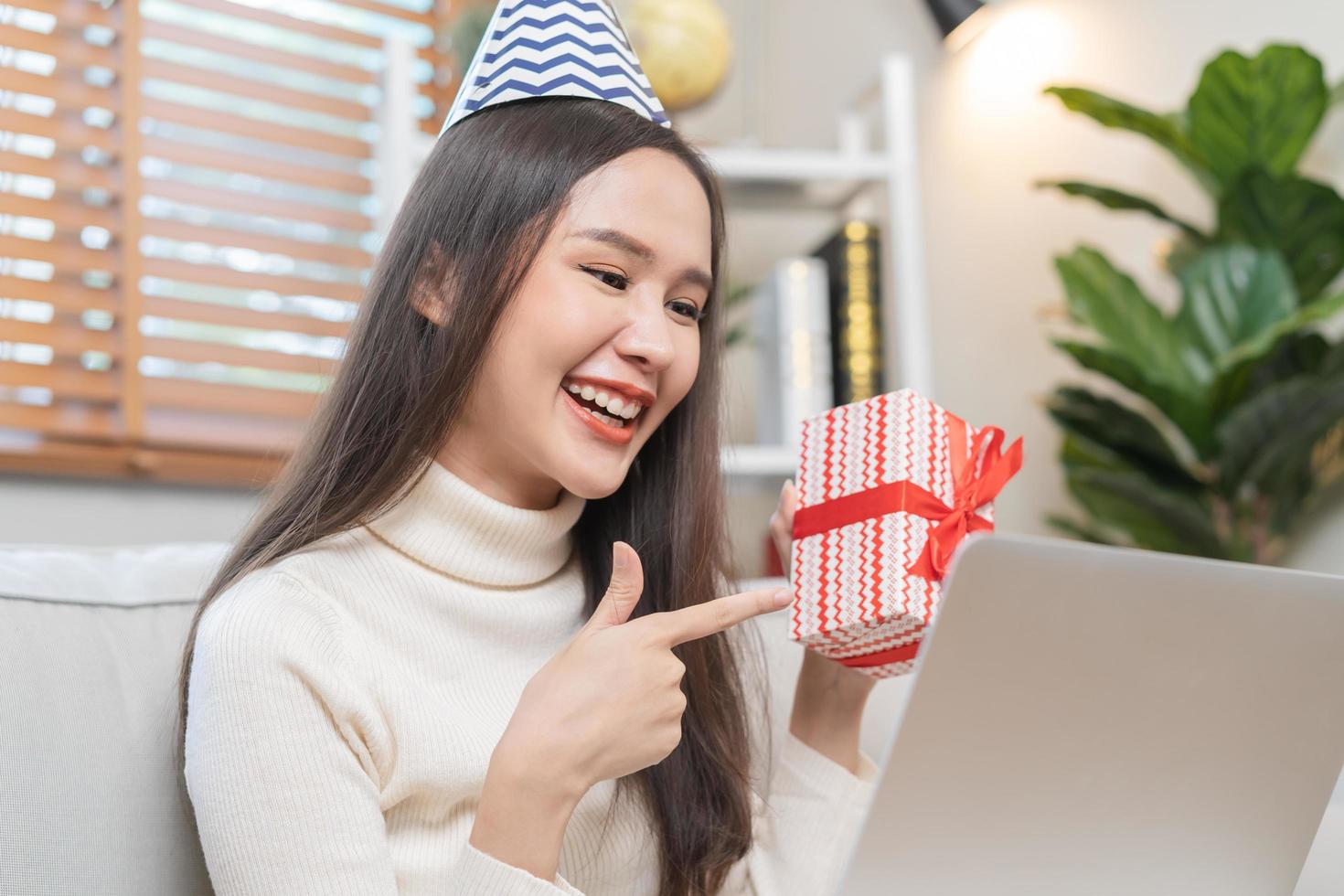 Celebrate on day of happy birthday season, pretty asian young woman using laptop computer, greeting on video call during social distance, holding gift box, happy to get present on couch, sofa at home. photo