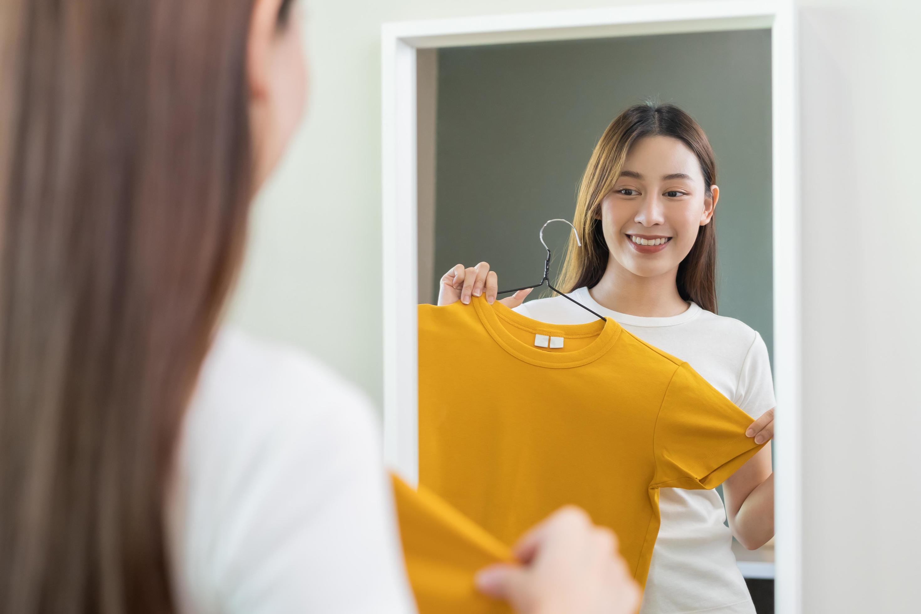 Choice of clothes,Nothing to wear. Attractive asian young woman, girl  looking into mirror, try on appare, choosing dress, outfit on hanger in  wardrobe at home. Deciding blouse what to put on which