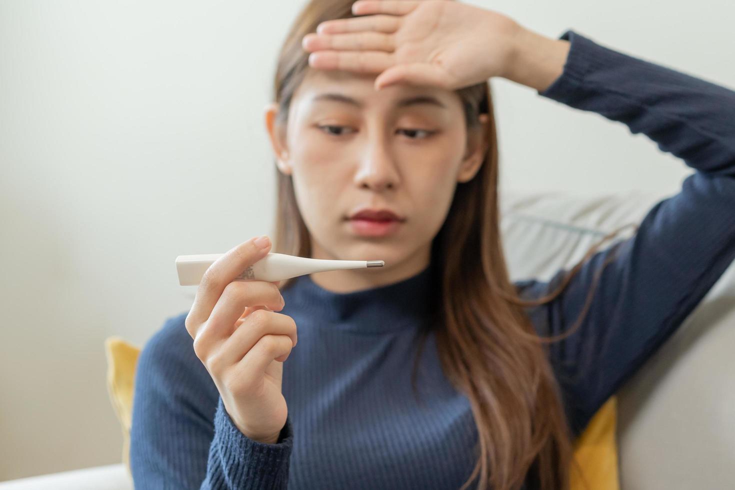 enfermo, influenza asiático joven mujer, niña dolor de cabeza tener un fiebre, gripe y cheque termómetro medida cuerpo temperatura, sensación enfermedad sentado en sofá cama a hogar. salud cuidado persona en virus, COVID-19. foto