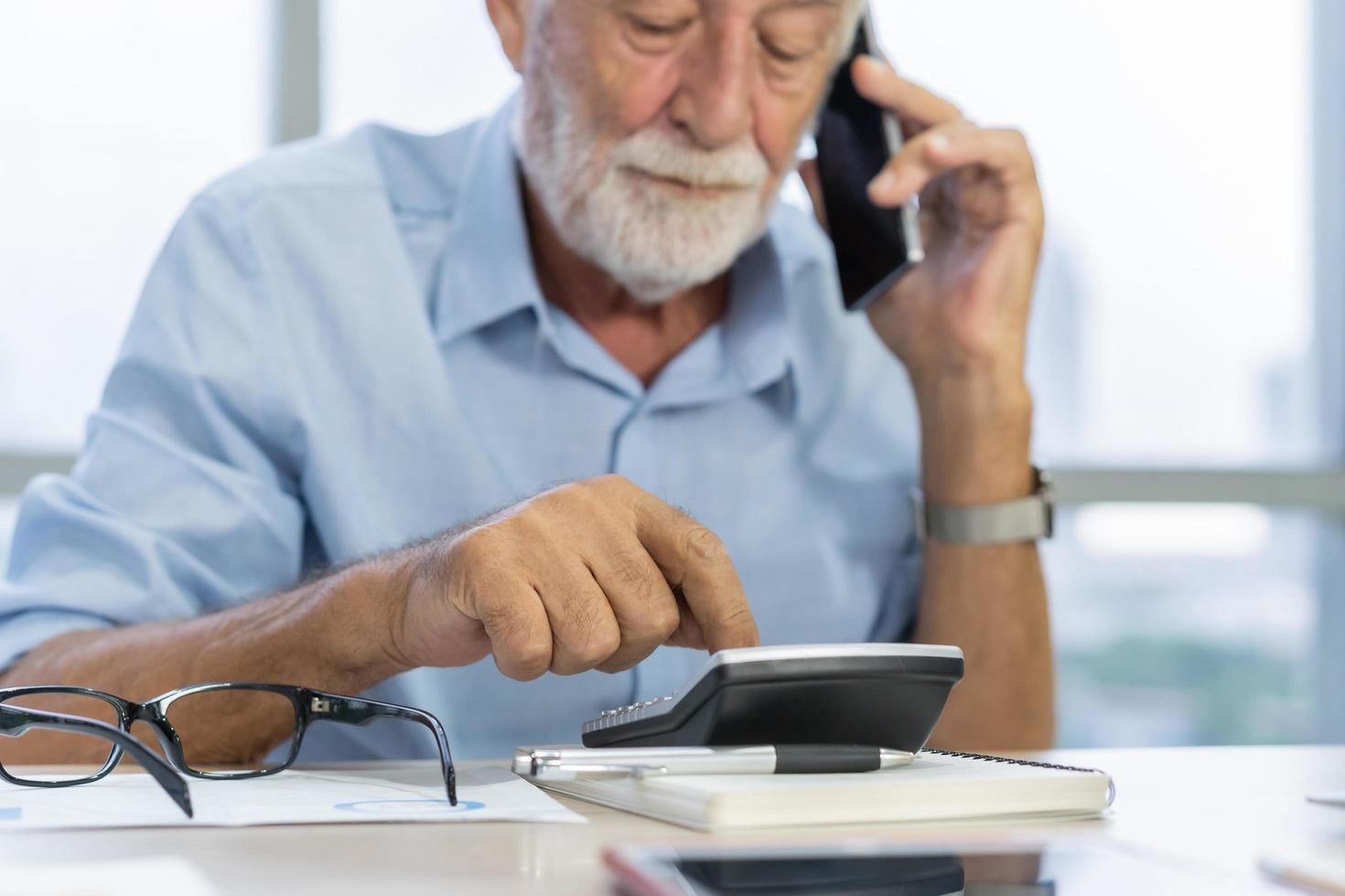 Business man of age senior, old caucasian mature, adult retired male calculate budget,  pay in deadline, talking mobile, phone call, using smartphone while sitting work at home office. Elderly worker. photo