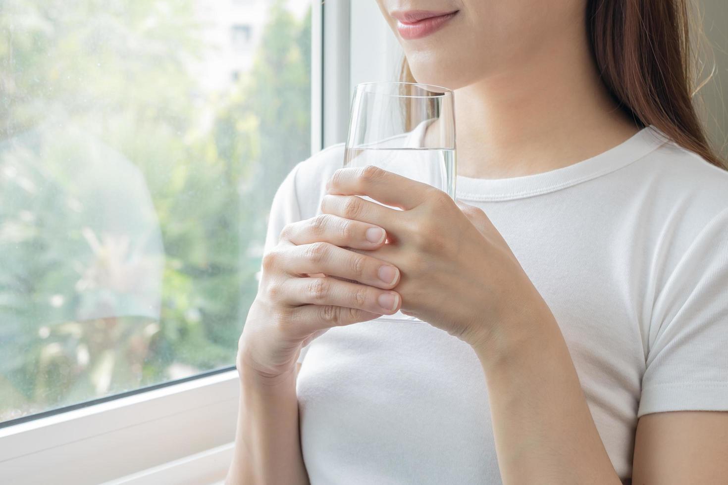 Happy beautiful, asian young woman, girl drinking, sip fresh glass of water for hydration of body, holding transparent glass in her hand, thirsty at home. Health care, healthy lifestyle concept. photo