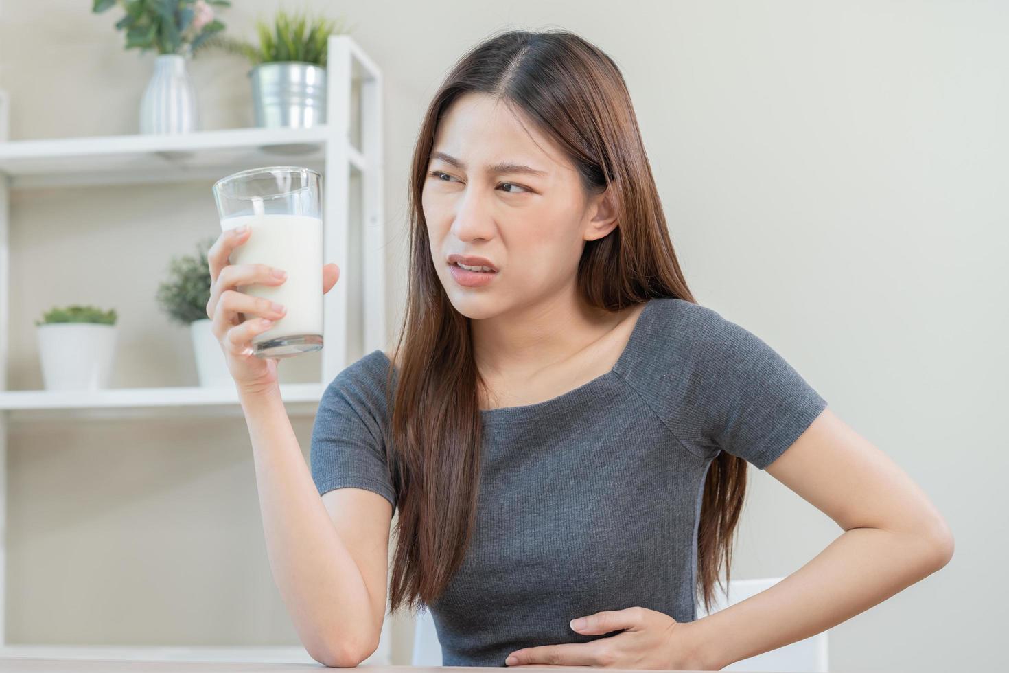 dolor, alergia asiático joven mujer, niña mano en barriga sufrir después bebida vaso de leche, infeliz y malo estómago dolor. lactosa intolerancia y lechería comida , salud problema concepto. foto