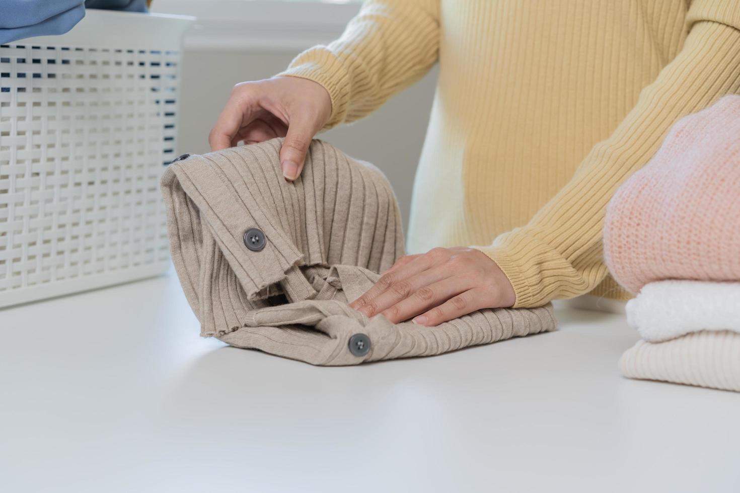 Housewife, asian young woman hand in many folding freshly shirts, sweaters or dress on desk, table after washing clean clothes and drying, making household working in room at home. Laundry and maid. photo