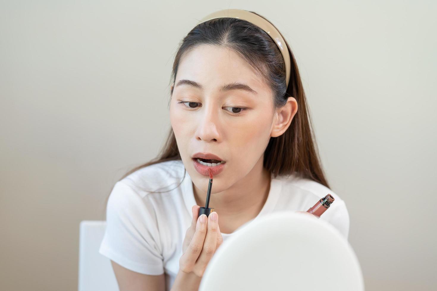 contento belleza bloguero, linda asiático joven mujer, niña mirando a espejo, maquillaje cara mano en aplicando lápiz labial, poniendo labio brillo en su boca a hogar. personas Mira con natural Moda estilo, Copiar espacio. foto