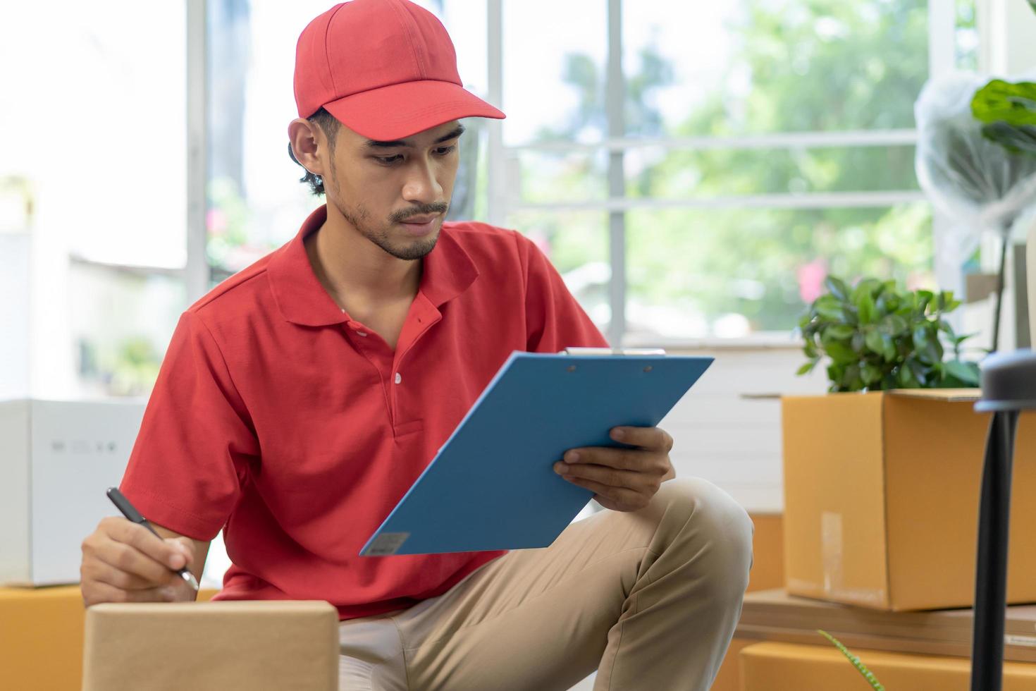 servicio, entrega asiático hombre, masculino comprobación lista cosa, paquete o empaquetar o cartulina caja, caja de cartón en almacén antes de entregar a cliente DIRECCIÓN. mensajero trabajando en uniforme. Moviente en nuevo reubicación foto