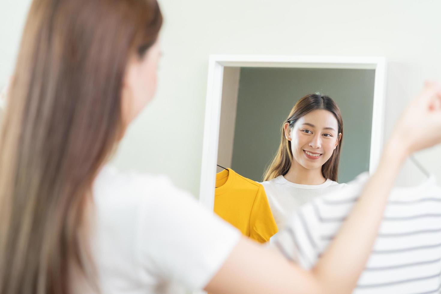 Choice of clothes,Nothing to wear. Attractive asian young woman, girl looking into mirror, try on appare, choosing dress, outfit on hanger in wardrobe at home. Deciding blouse what to put on which one photo