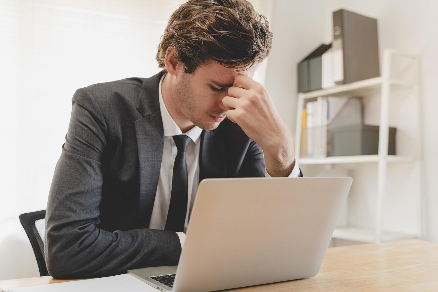 exhausto, estresado caucásico joven negocio hombre es trabajando en negro traje, masaje nariz desde seco ojos, sufrir en difícil trabajo mientras utilizar ordenador portátil computadora a oficina hogar. tiempo extraordinario trabajo, deuda problema gente. foto