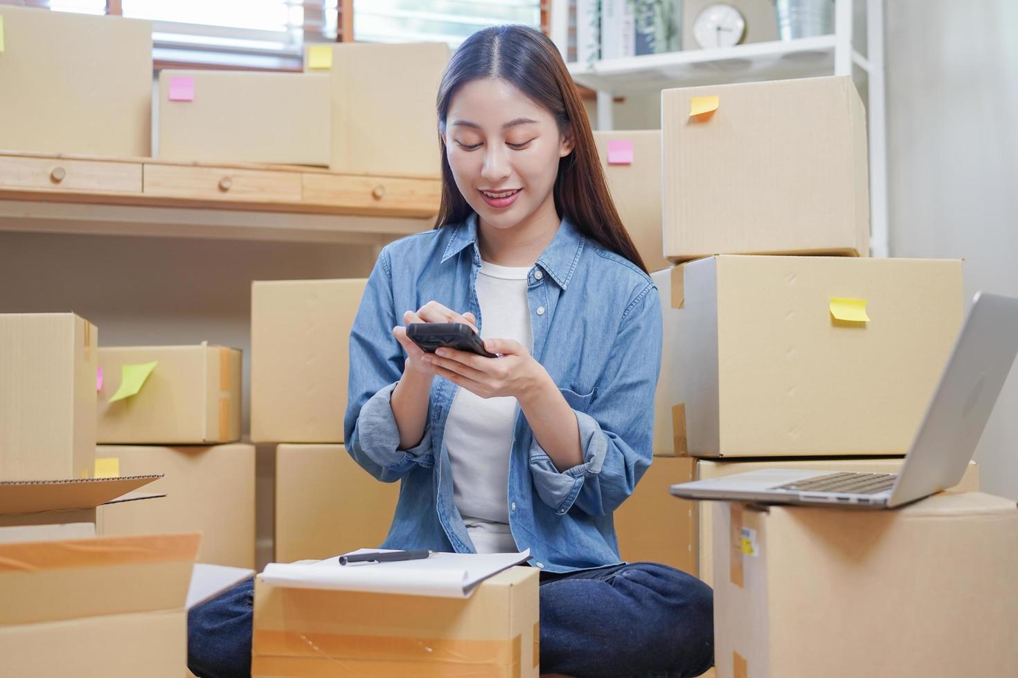 Small business entrepreneur SME, asian young woman owner packing product, checking parcel box delivery, using smartphone receive order from customer, working at home office. Merchant online, ecommerce photo