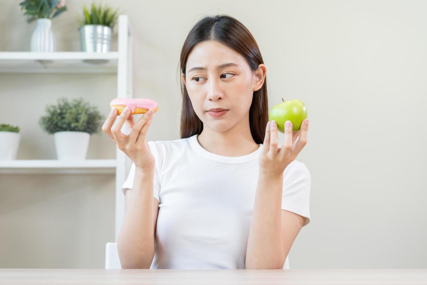 Diet, Dieting asian young woman or girl smile, confused choose, choice green apple or pink sweet donut, at home, eat food for good healthy, health when hungry. Close up female weight loss person. photo