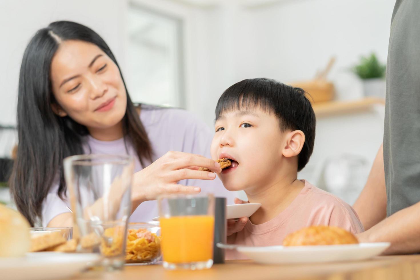 contento refresco familia desayuno en mañana, asiático joven padre padre, madre y pequeño linda chico, niño teniendo comida en cocina comiendo juntos a hogar. alegre, disfrutar Cocinando gente. foto