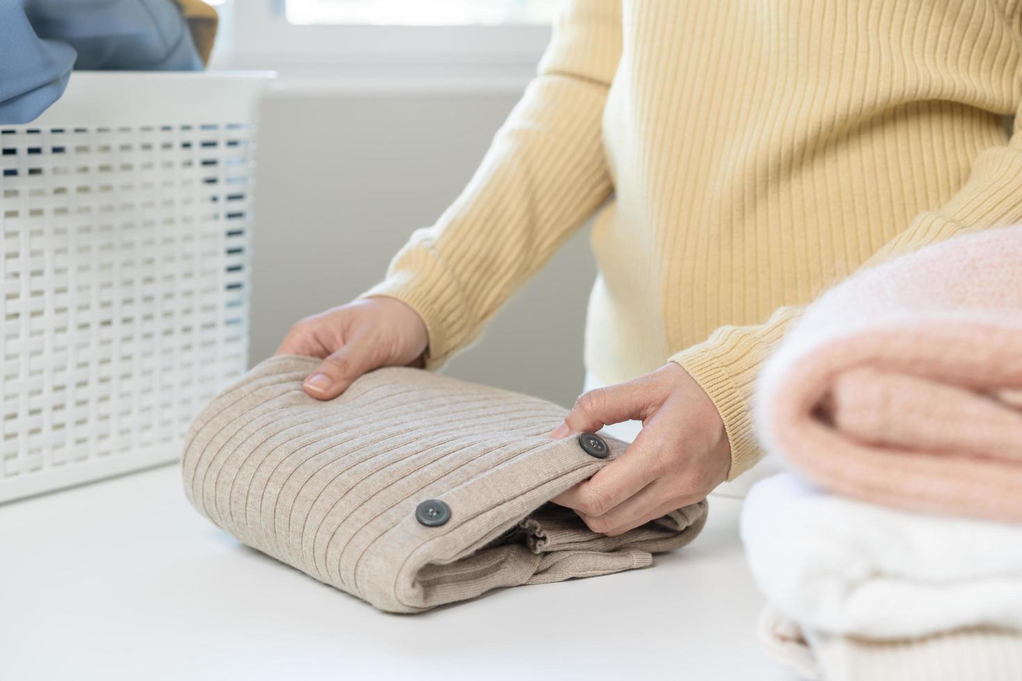 Housewife, asian young woman hand in many folding freshly shirts, sweaters or dress on desk, table after washing clean clothes and drying, making household working in room at home. Laundry and maid. photo