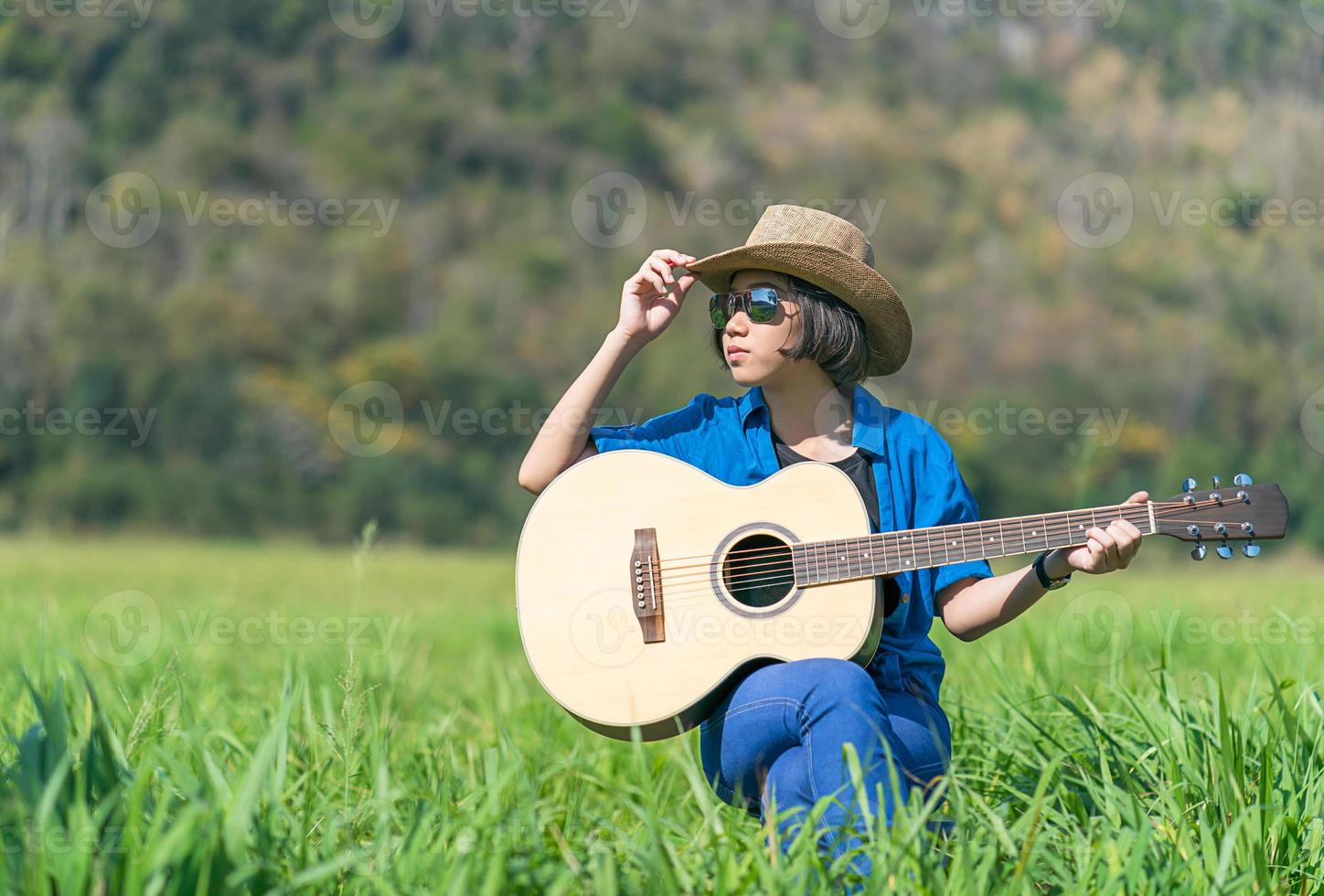 Women short hair wear hat and sunglasses sit playing guitar in grass field photo