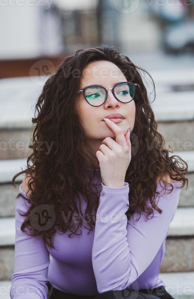 Beautiful young woman with brunette curly hair, portrait in eye glasses enjoying the sun in the city. photo