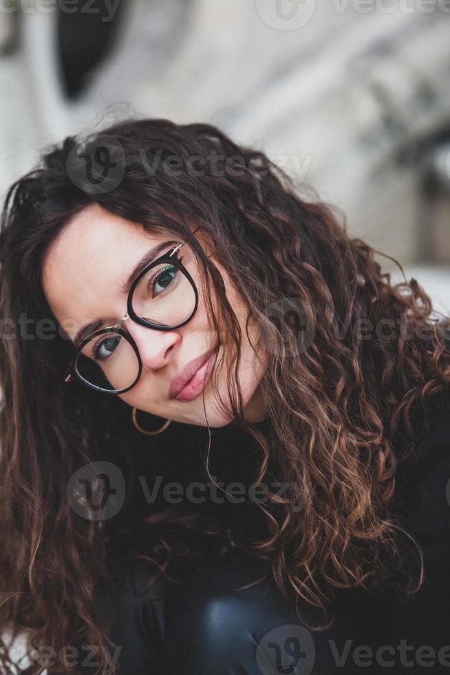 Beautiful young woman with brunette curly hair, portrait in eye glasses enjoying the sun in the city. photo