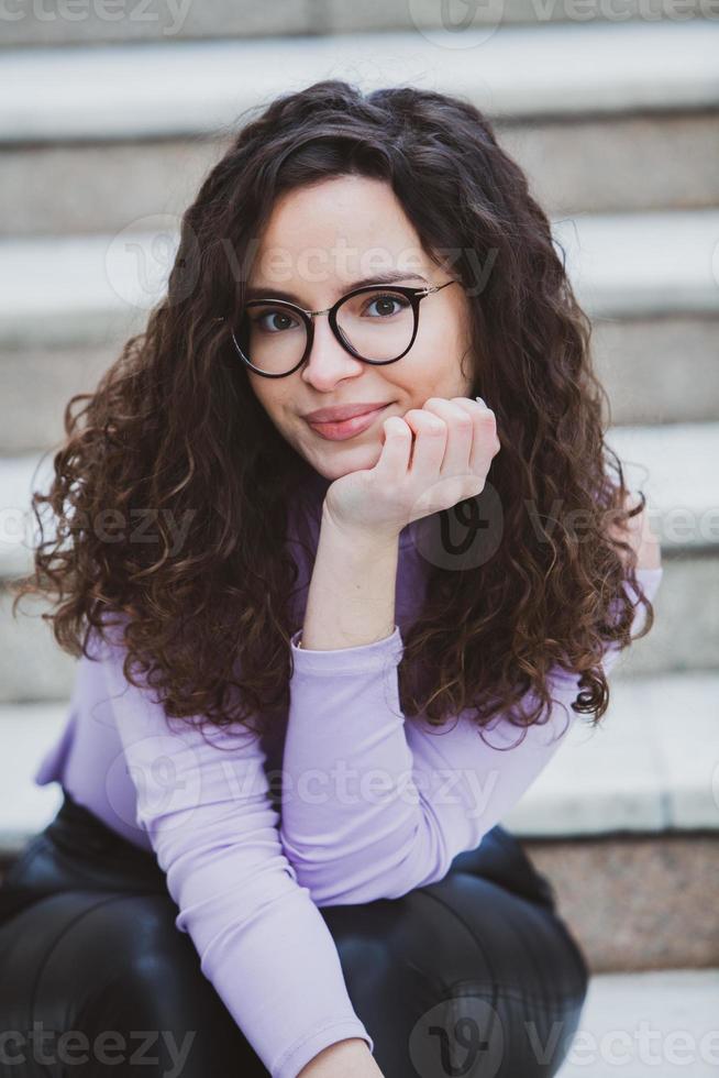 Beautiful young woman with brunette curly hair, portrait in eye glasses enjoying the sun in the city. photo