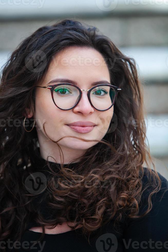 Beautiful young woman with brunette curly hair, portrait in eye glasses enjoying the sun in the city. photo