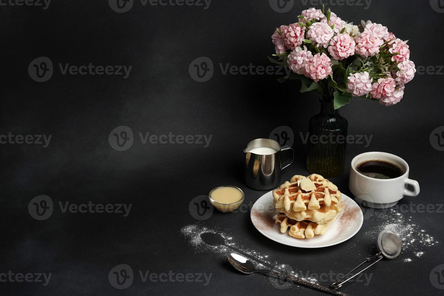 Tasty Waffles Plate, Caramel Sauce, Coffee Cup, Milk, dessertspoon, strainer, pink flowers on a Black concrete Background photo