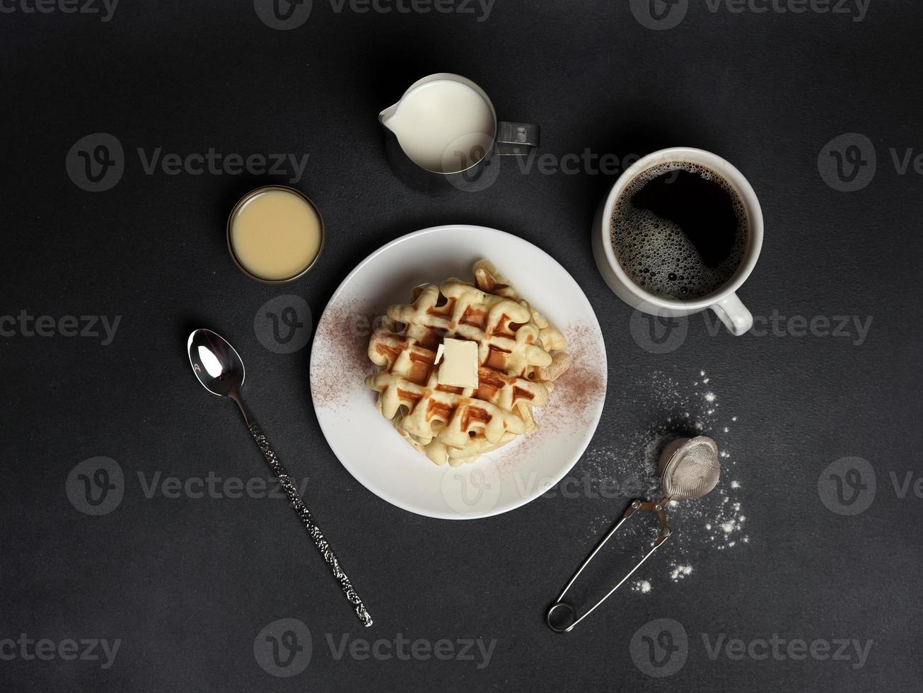 Top view of tasty Waffles Plate, Caramel Sauce, Coffee Cup, Milk, dessertspoon, strainer on a Black concrete Background photo