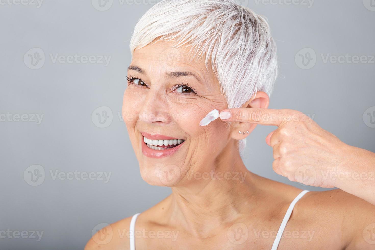 sonriente mayor mujer aplicando anti-envejecimiento loción a eliminar oscuro círculos debajo ojos. maduro mujer utilizando cosmético crema a esconder arrugas dama utilizando día hidratante a contrarrestar el envejecimiento de el piel. foto