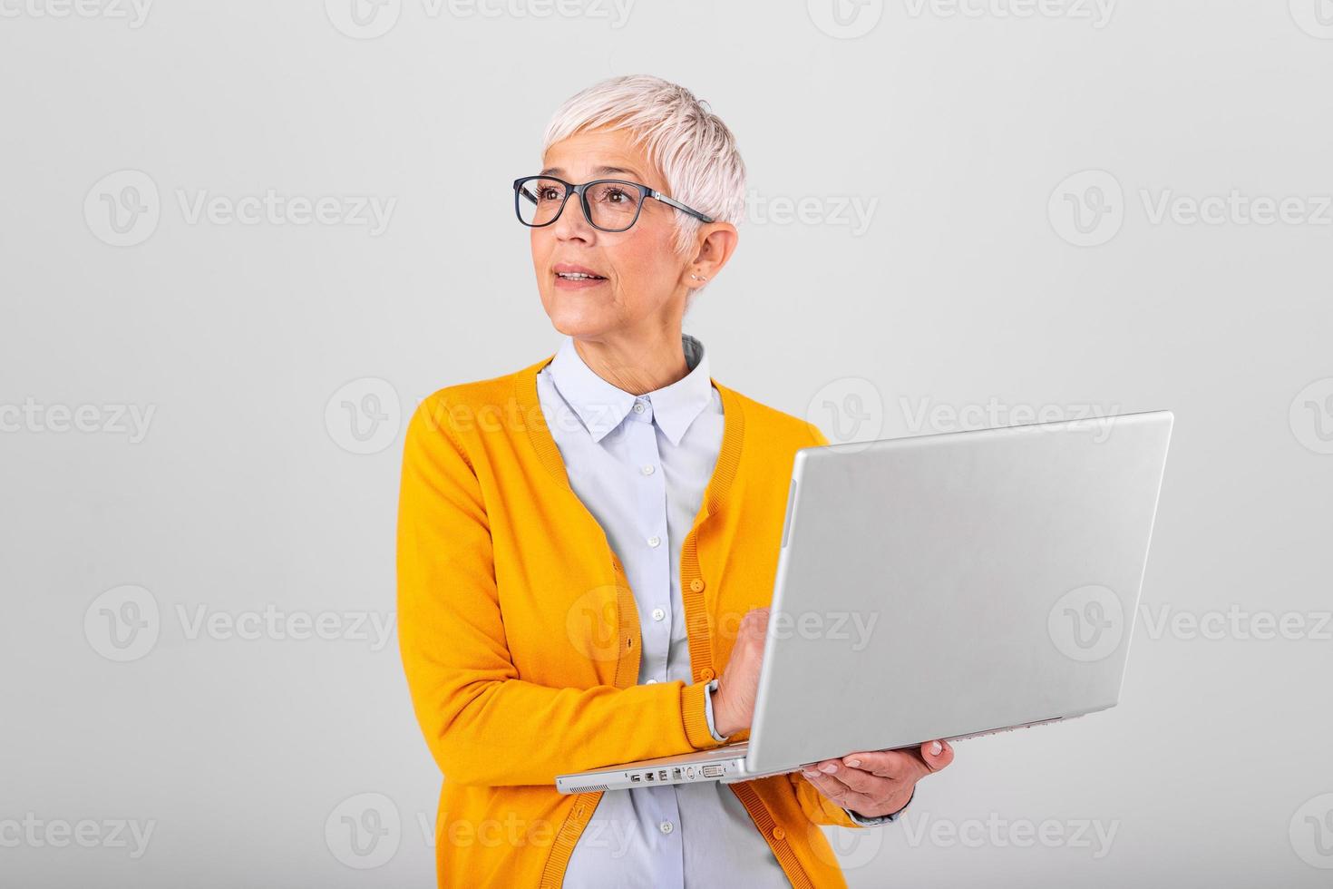 Photo of a thinking mature business woman isolated over grey background holding laptop computer. Image of confused senior woman using laptop computer. Looking at laptop while holding face with hands.