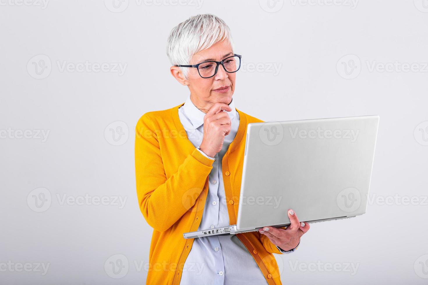 Photo of a thinking mature business woman isolated over grey background holding laptop computer. Image of confused senior woman using laptop computer. Looking at laptop while holding face with hands.