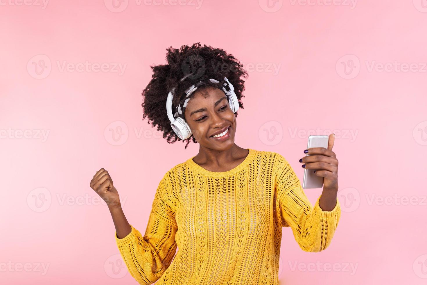 estilo de vida concepto - retrato de hermosa africano americano mujer alegre escuchando a música en móvil teléfono. de moda elegante linda niña en auriculares escuchando a música bailando aislado rosado antecedentes foto