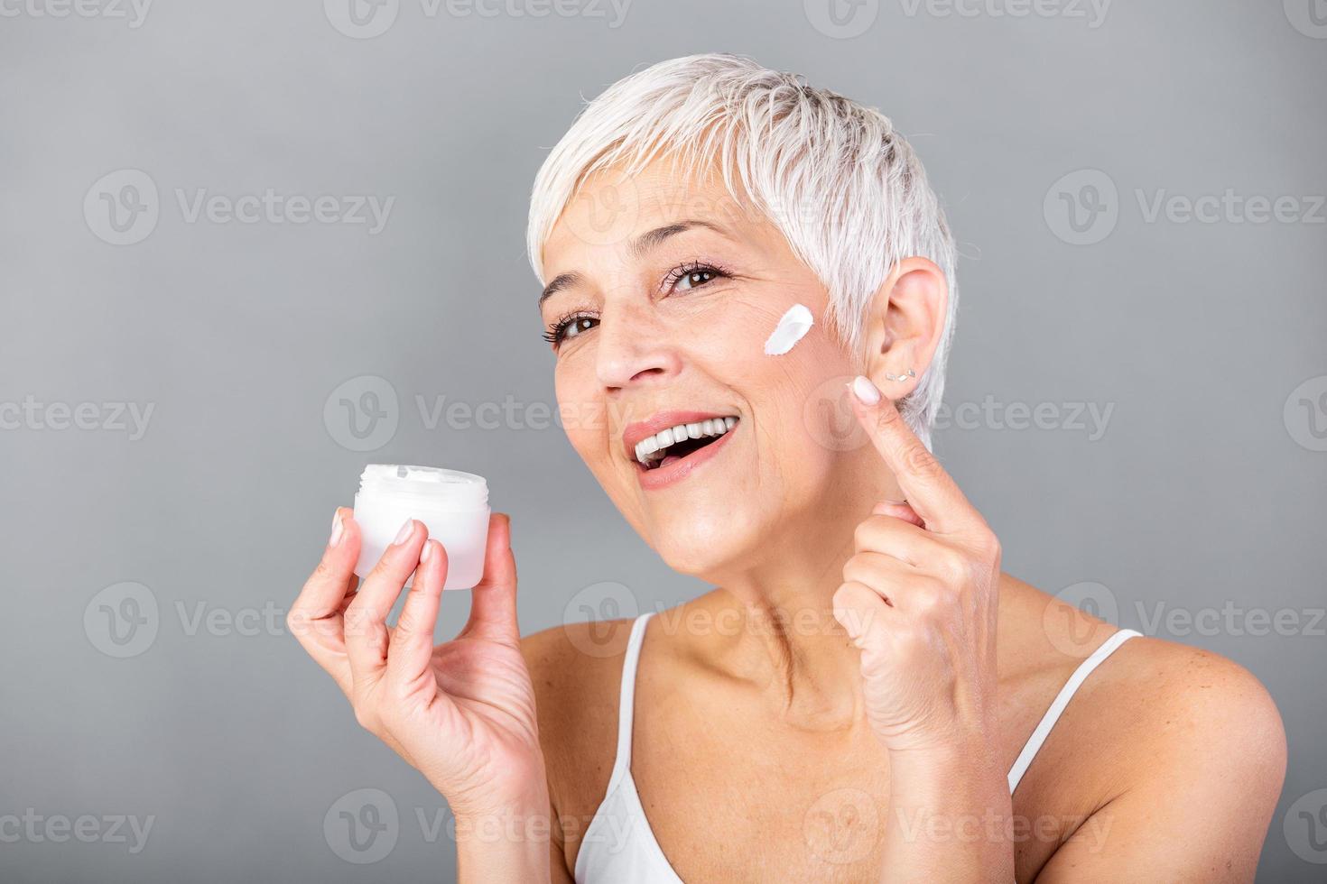 Beautiful mature woman holding jar of moisturizer and looking at camera. Happy senior woman holding a bottle of anti-aging lotion isolated over grey background. Anti aging and beauty treatment. photo