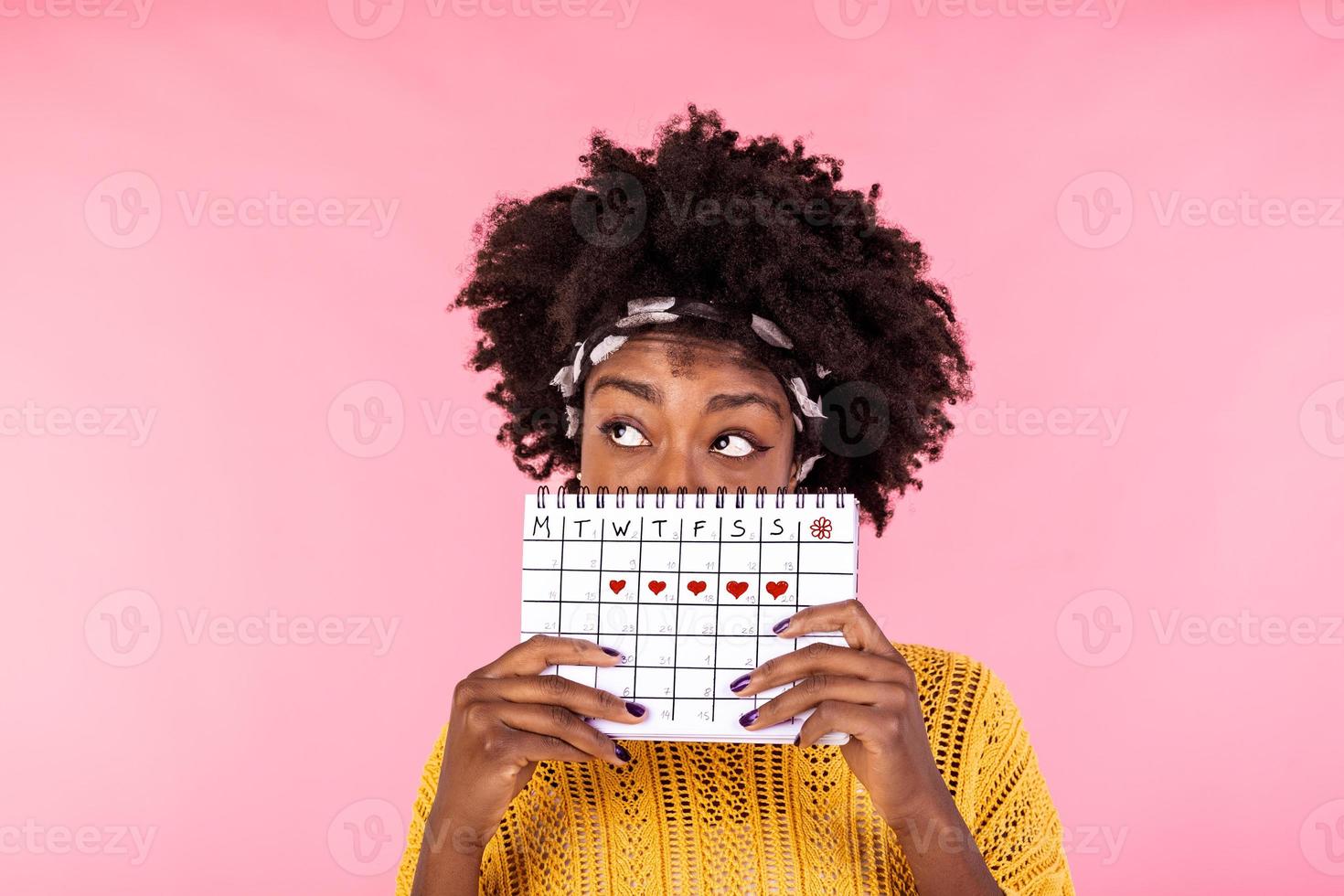 Portrait of a funny young African American girl in hiding behind a menstrual periods calendar and looking away at copy space isolated over pink background. Female Period calendar photo