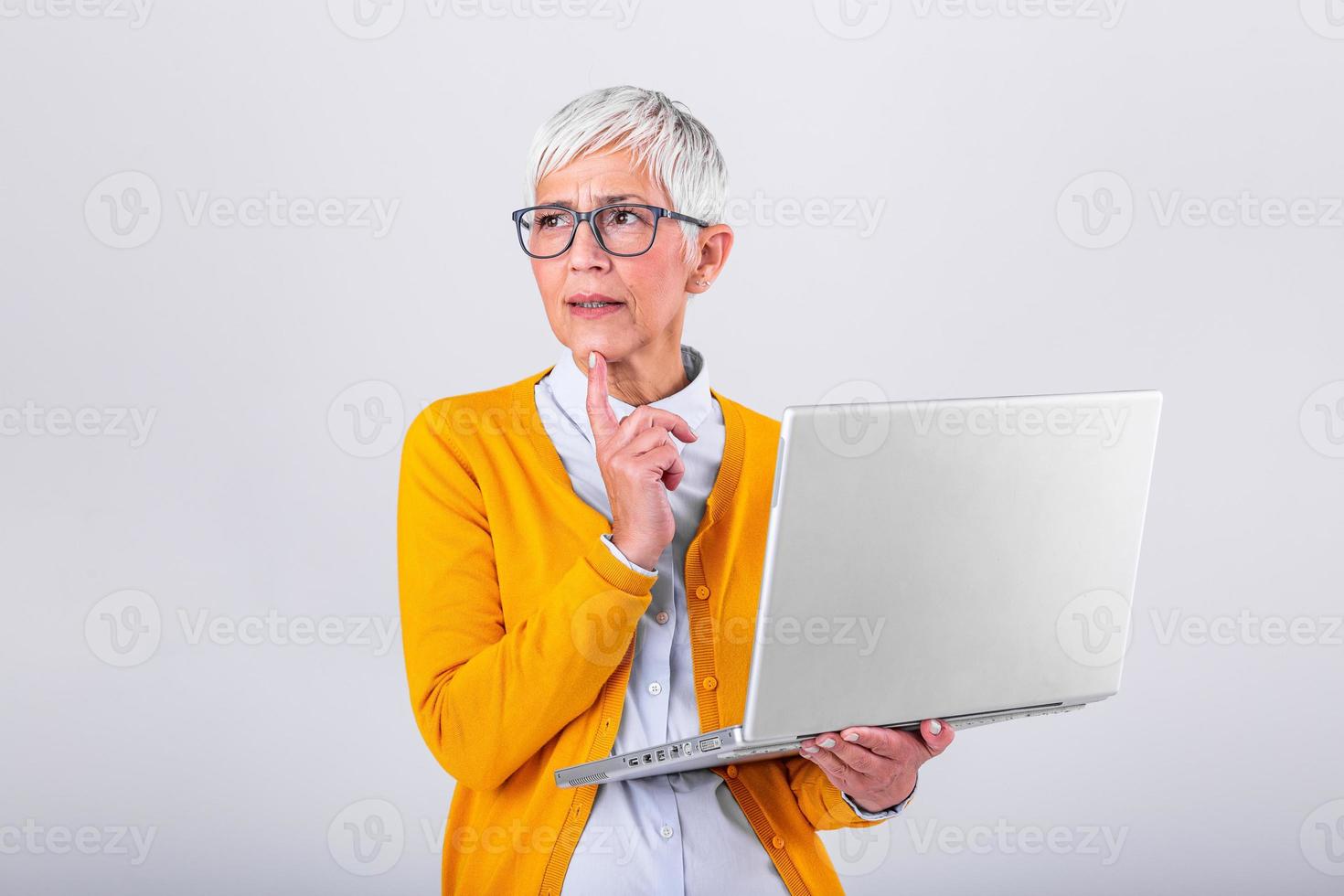 Photo of a thinking mature business woman isolated over grey background holding laptop computer. Image of confused senior woman using laptop computer. Looking at laptop while holding face with hands.