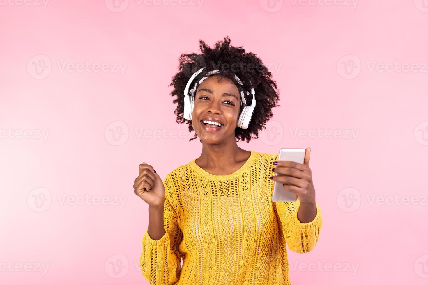 estilo de vida concepto - retrato de hermosa africano americano mujer alegre escuchando a música en móvil teléfono. de moda elegante linda niña en auriculares escuchando a música bailando aislado rosado antecedentes foto