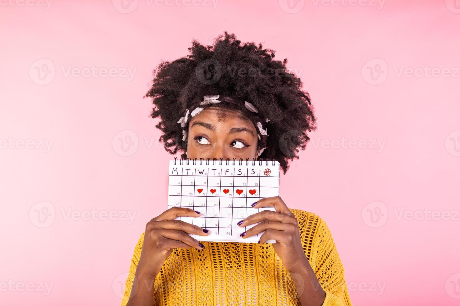 Portrait of a funny young African American girl in hiding behind a menstrual periods calendar and looking away at copy space isolated over pink background. Female Period calendar photo