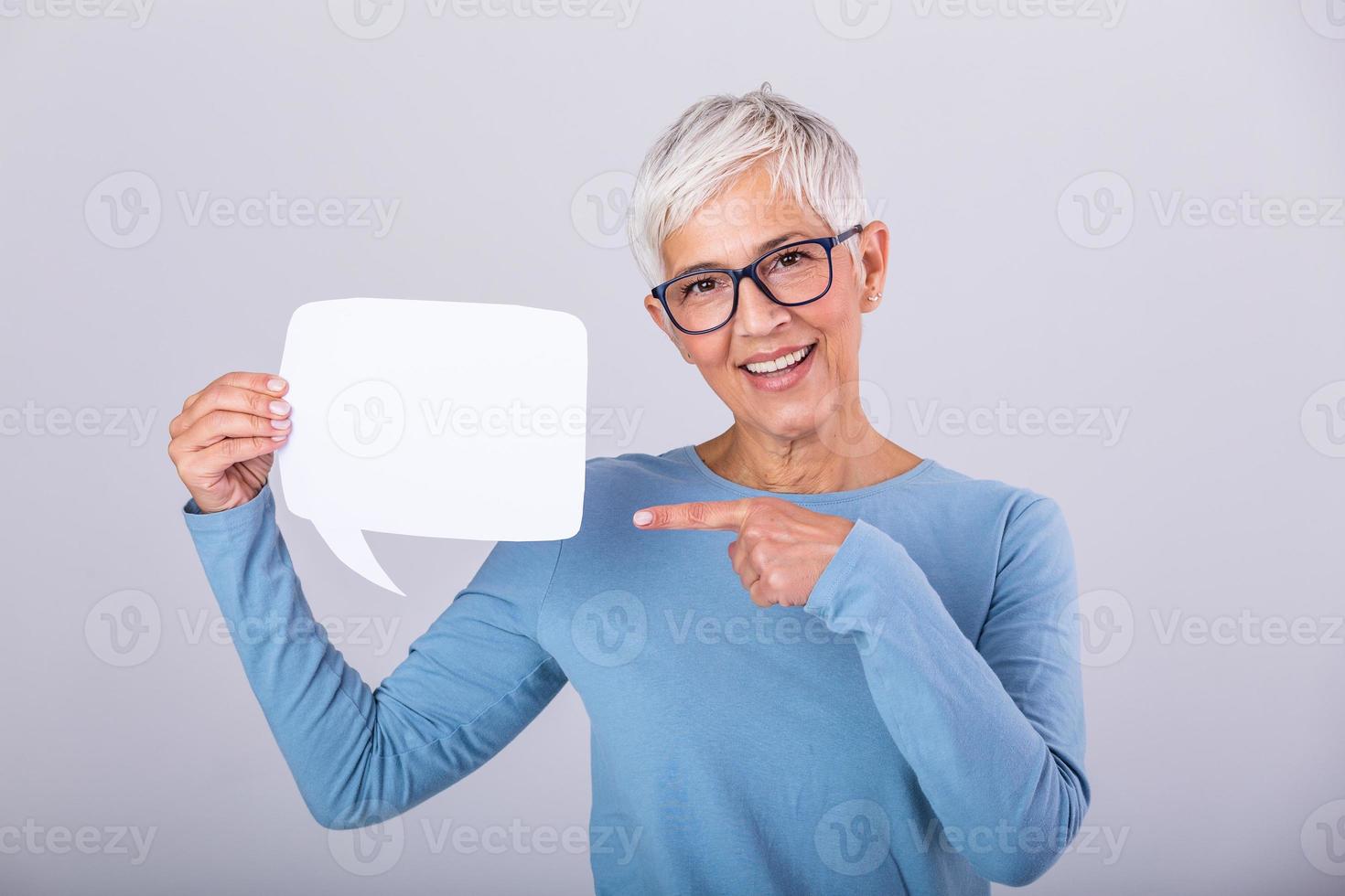Happy mature woman in plain blue long sleeve t-shirt holding empty speech bubble isolated on background. Woman showing sign speech bubble banner looking happy and pointing her finger on it photo